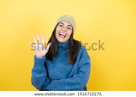 Young beautiful woman wearing blue casual sweater and wool hat showing and pointing up with fingers number five while smiling confident and happy