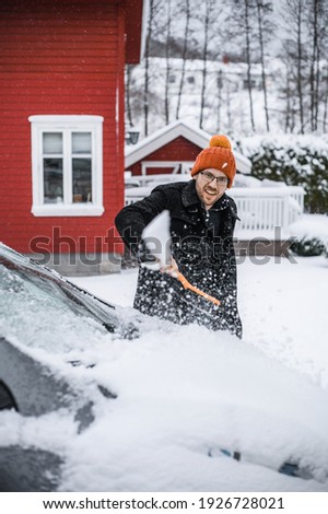 grey   electric car with Scandinavian house in the background. A nerdy guy with brush is removing snow.