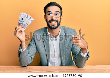 Young hispanic man holding colombian pesos banknotes sitting on the table smiling happy and positive, thumb up doing excellent and approval sign 
