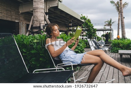 Side view of attractive barefoot female in summer clothes sitting on chair and reading ebook while spending time on terrace of tropical resort