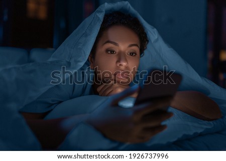 technology, internet, communication and people concept - young african american woman with smartphone lying under blanket in bed at home at night Royalty-Free Stock Photo #1926737996
