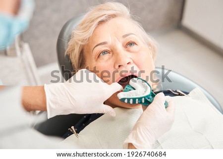 Cropped view of the mature blonde haired woman sitting with open mouth while her dentist putting at her mouth dental impression for denture. Stomatology concept