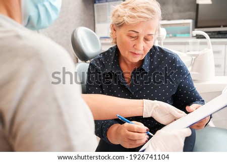 New smile. Happy senior female patient sitting in front of her dentist and smiling while being thankful after her dental treatment. Woman signing th documents at the dental clinic Royalty-Free Stock Photo #1926741104