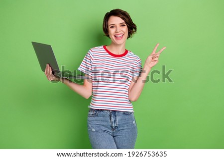 Portrait of attractive cheerful girl holding laptop showing v-sign having fun isolated over green pastel color background