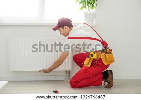 Man in work overalls using wrench while installing heating radiator in room. Young plumber installing heating system in apartment. Concept of radiator installation, plumbing works and home renovation Royalty-Free Stock Photo #1926766487