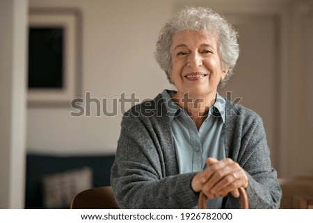 Portrait of smiling mature woman holding walking stick and sitting on chair at home. Portrait of happy senior woman under quarantine during covid-19 pandemic smiling while looking at camera. Royalty-Free Stock Photo #1926782000