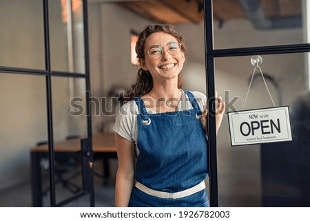 Portrait of positive business woman standing at cafeteria door entrance. Cheerful young waitress in blue apron near glass door with open signboard and looking at camera. Excited small business owner. Royalty-Free Stock Photo #1926782003