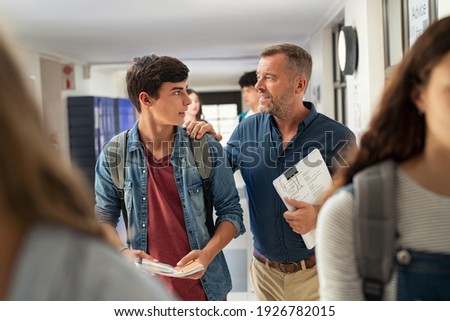 Mature man talking to high school student in hallway at the end of the lesson. Senior professor in conversation with guy student in campus. Teacher talking to encourage young man after exam. Royalty-Free Stock Photo #1926782015
