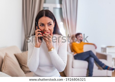 Young woman Talking Privately On Cell Phone While Her husband is watching tv in the back. Beautiful young woman cheating on boyfriend talking with lover