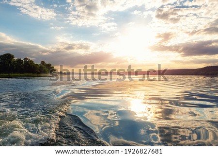 Scenic bright vibrant blue to red warm sunset evening time landscape with motorboat swirl trace on water surface of lake or river. Horizon coast line on background. Scenic fog haze near ground