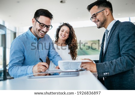 Middle age couple choosing and buying car at car showroom. Car salesman helps them to make right decision. Man signs buyers contract.