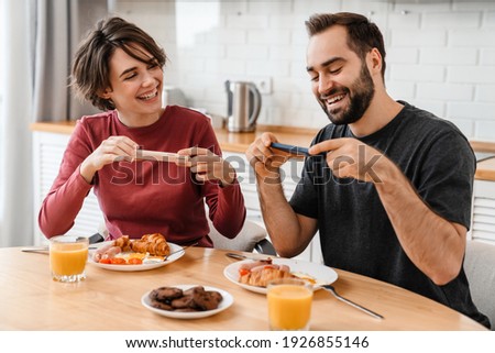 Portrait of young caucasian couple taking photo on cellphone while having breakfast in cozy kitchen at home
