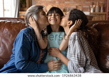 Portrait of happy little Latino 7s girl child kissed by young mom and senior grandmother, relax together at home. Loving mother and mature grandparent congratulate excited small kid. Family concept.