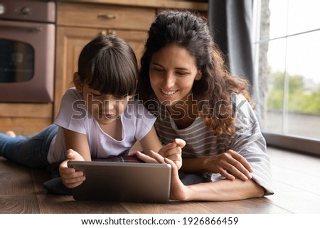 Caring young Hispanic mom and little daughter use modern tablet watch video online together. Happy Latino mother and small girl child look at pad screen study or play on device on internet at home.
