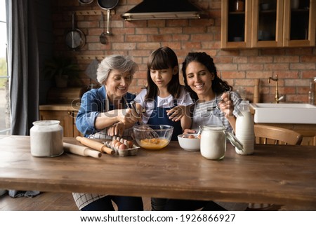 Happy three generations of Hispanic women gather in kitchen cook delicious breakfast together. Smiling little girl with young mom and mature grandmother prepare pancakes or bake at home on weekend. Royalty-Free Stock Photo #1926866927