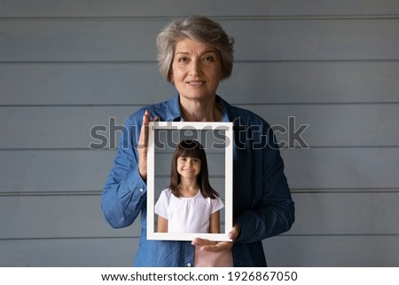 Portrait of smiling mature Hispanic grandmother isolated on grey wall background show granddaughter picture in frame. Elderly senior woman hold photo of her photo as child. Now and then concept.