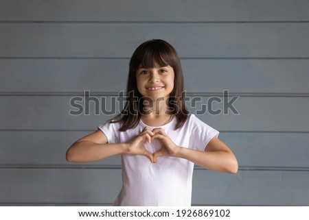 Portrait of smiling Hispanic little girl kid isolated on grey wall background show hear love hand gesture or sign. Happy Latino small 8s kid feel grateful thankful demonstrate care. Charity concept.
