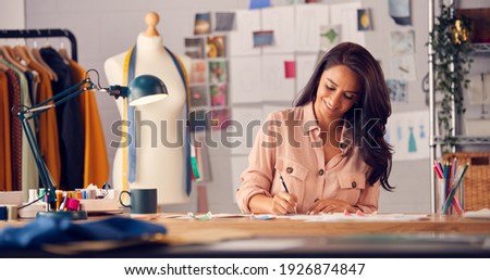 Female Fashion Designer In Studio Working On Sketches And Designs At Desk