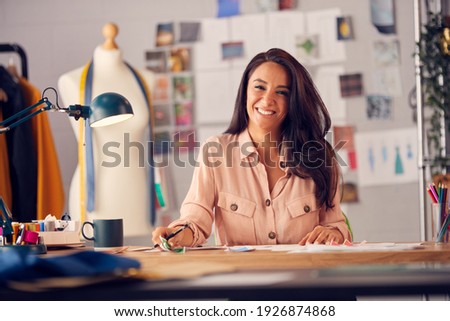 Portrait Of Female Fashion Designer In Studio Working On Sketches And Designs At Desk