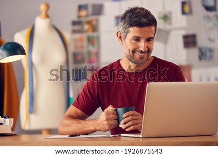 Mature Male Fashion Designer In Studio Sitting At Desk Working On Laptop