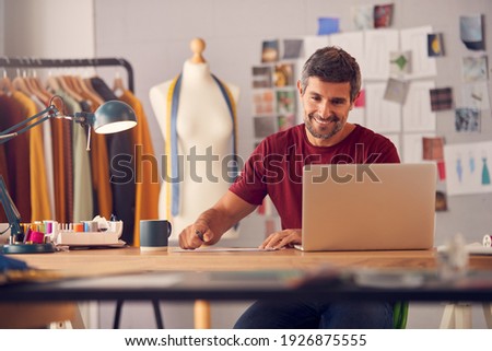 Mature Male Fashion Designer In Studio Working On Sketches Or Documents At Desk With Laptop