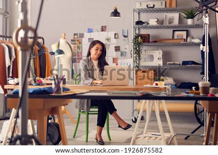 Female Fashion Designer In Studio Working On Sketches Or Documents At Desk With Laptop