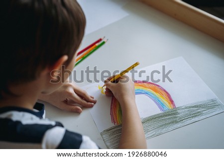 adorable caucasian boy of elementary age drawing a rainbow with pencils sitting at the desk in his room at home. View from behind his shoulder.Image with selective focus