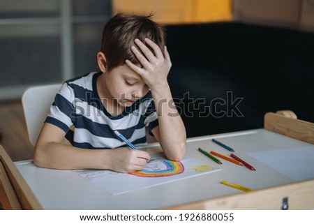 adorable caucasian boy of elementary age drawing a rainbow with pencils sitting at the desk in his room with head reclined upon his hand. Image with selective focus