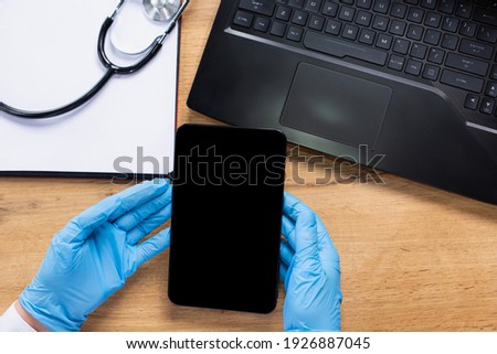 Hands of a paramedic in latex gloves holding a tablet. On the table a laptop, paper and a stethoscope. Cropped photo.