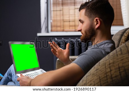 Man sitting relaxed on a couch works on a laptop with empty green screen. Dayout in his living room man uses notebook computer. Office online work
