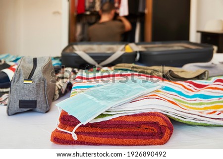 an open suitcase on the bed, packed with different clothes, such as swim trunks, beach towels, and some blue disposable face masks, and a young caucasian man searching in his closet in the background