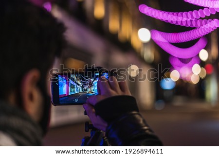 A young man is taking a picture with his smartphone on a tripod. He is wearing a face mask. In the phone we can see an empty street.