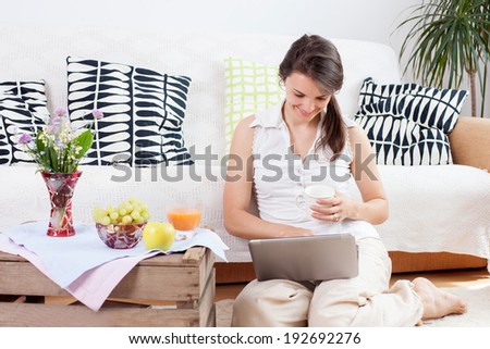 Young woman, sitting on the floor, reading from computer and eating healthy breakfast