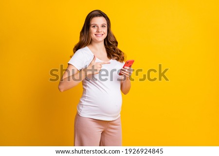 Photo of pretty lady waiting baby dressed white clothes tummy pointing modern device empty space isolated yellow color background