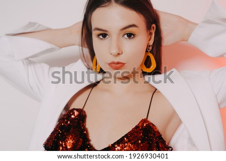 A gorgeous brunette in a shiny red dress is dancing, a model in a white jacket is having fun. Girl in yellow accessories, earrings and make-up on a pink background.