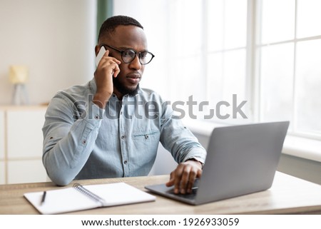 Serious Black Businessman Talking On Mobile Phone Using Laptop Computer Sitting At Workplace In Modern Office. Business Communication And Negotiation Via Cellphone Concept Royalty-Free Stock Photo #1926933059