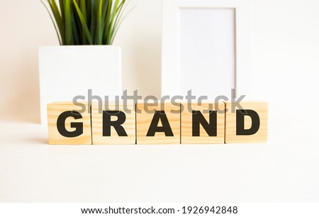 Wooden cubes with letters on a white table. The word is GRAND. White background with photo frame, house plant.