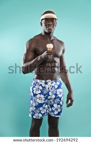 Summer black african american muscled fitness man holding ice cream. Wearing blue swimming shorts and cap. Studio shot against blue.