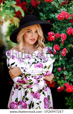Outdoor photo of romantic young woman in rose garden. girl with red lips in a dress with a print of roses. Young model in hat on a background of a bush of roses. Stylish woman.