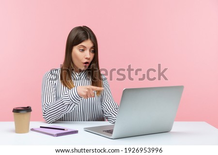 Wow, unbelievable! Surprised excited woman employee pointing at laptop screen, reading shocking news or talking on video call with amazed expression. Indoor studio shot isolated on pink background