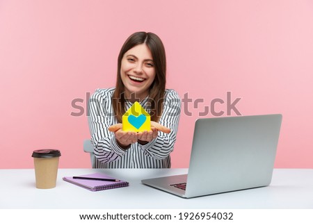 Smiling positive woman real estate agent holding model of paper house with heart in hands sitting at workplace with laptop, safety living construction. Indoor studio shot isolated on pink background