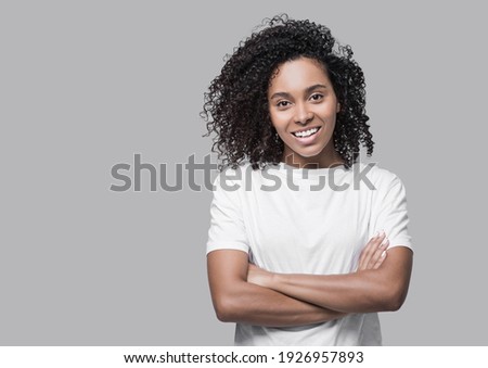 Studio portrait of a beautiful young woman with black curly hair. African american girl with crossed arms looking at camera. Isolated on grey background. People, lifestyle, beauty concept