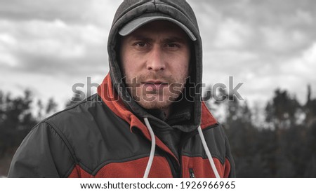 Portrait of a man in a cap and hood with bristles on his face. A guy with a serious face on the background of the forest