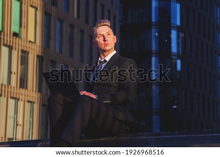 Young man businessman in style wear clothes, suit sitting on steps with laptop evening on sunset after work. Relaxed male banker in background office located in financial district. Copy space