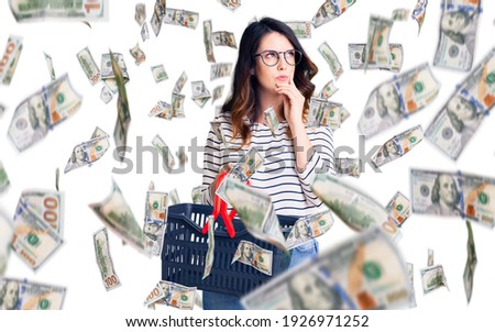 Beautiful young brunette woman holding supermarket shopping basket serious face thinking about question with hand on chin, thoughtful about confusing idea