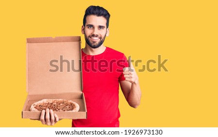 Young handsome man with beard holding delivery cardoboard with italian pizza smiling happy and positive, thumb up doing excellent and approval sign 