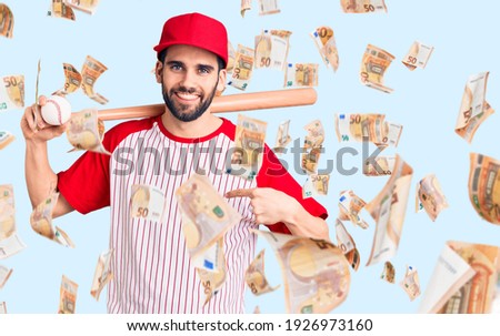 Young handsome man with beard playing baseball holding bat and ball pointing finger to one self smiling happy and proud