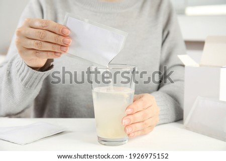 Woman pouring powder from medicine sachet into glass with water at table, closeup