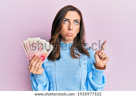 Young brunette woman holding 20 israel shekels banknotes pointing up looking sad and upset, indicating direction with fingers, unhappy and depressed. 