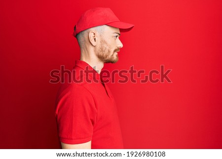 Young redhead man wearing delivery uniform and cap looking to side, relax profile pose with natural face with confident smile. 